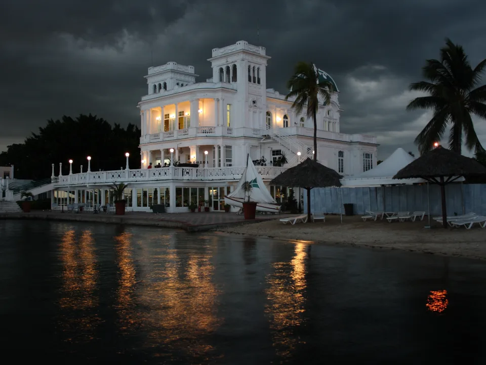 An illuminated waterfront building and palms reflected in the water at dusk in Cienfuegos, Cuba
