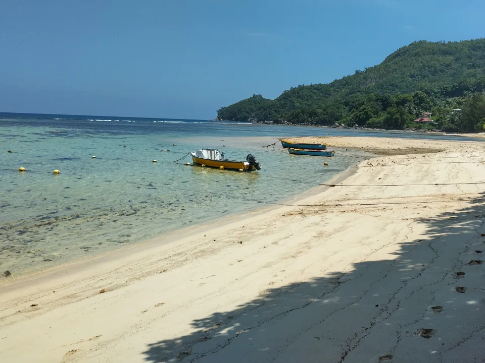 Takamaka Beach on Mahé in Seychelles, with turquoise shallows, small local boats, pale sand and a wooded headland